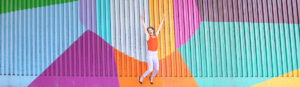 This is an image of Sarah Follent jumping for joy in front of a rainbow wall.