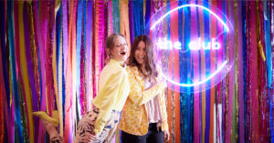 This is an image of Clarissa and Sarah dancing in front of a colourful streamer wall and Club neon light!